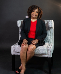 picture of Zelma McKinney smiling with curly brown hair, a pearl necklace, a red shirt, and a black jacket sitting in a gray chair
